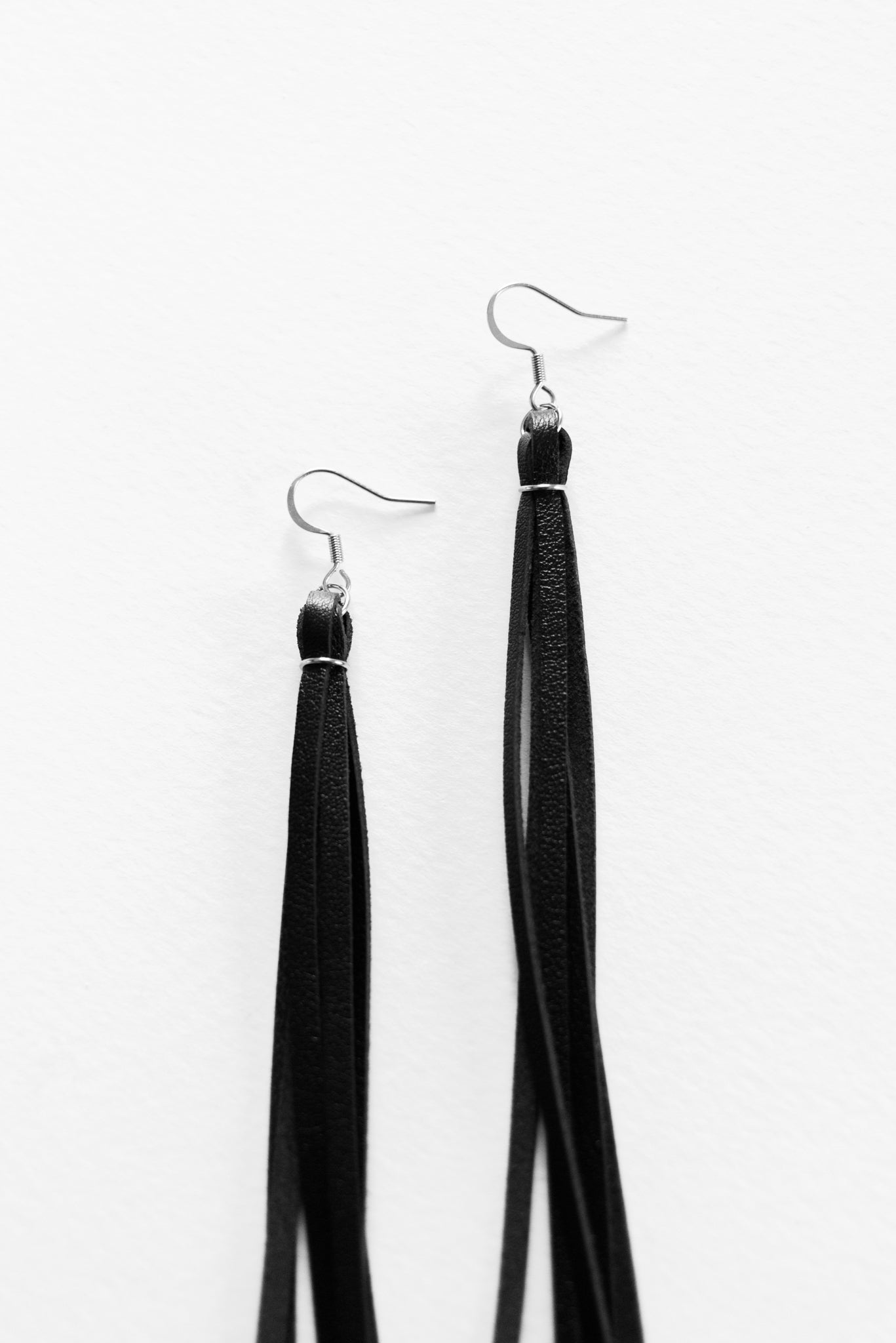 Close up view of silver french wire earrings with long black leather fringe, resting on a white backdrop